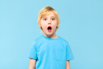 Wow! Portrait of a shocked cute little boy with blond hair on pastel blue background. Surprised preschooler studio shot, looking at camera.