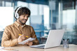 © Liubomir - Businessman is studying inside the office, man is upgrading his qualifications, watching online video course, recording data, worker in headphones, talking, using video call.
