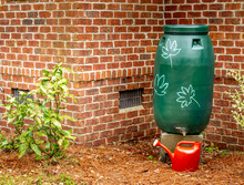 Green Rain Water Barrel Next To A Red Brick Wall With Watering Can, Sustainable Living