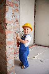 Wall Mural - Full length of happy child construction worker holding hammer tool and looking at camera while standing by brick wall at home during renovation. Smiling kid wearing safety helmet and work overalls.