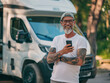© JW Studio - Mature tattooed man standing near rv camper van on vacation using mobile phone. Smiling mature active traveler holding smartphone enjoying free internet in camping tourism nature park