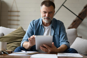 Cheerful senior man in casual clothing using digital tablet while sitting on the sofa at home.