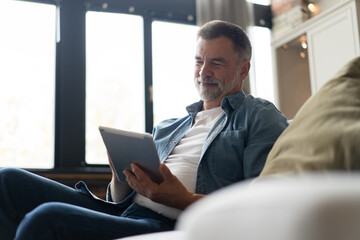 Cheerful senior man in casual clothing using digital tablet while sitting on the sofa at home.