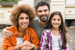 Cheerful mixed-race family of three parents and daughter looking at camera hugging together road trip adventure by motor home trailer caravanning by camper van