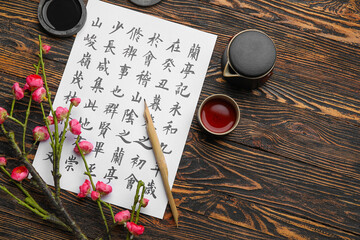 Wall Mural - Paper sheet with Asian hieroglyphs, nib pen, ink, cup of tea and blooming branch on wooden background. International Haiku Poetry Day
