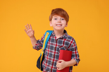 Sticker - Happy schoolboy with backpack and book waving hello on orange background