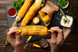 Grilled corn on the cob in caucasian hands on kitchen wooden table background