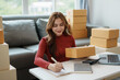 © amnaj - Smiling young business owner woman prepare parcel box and standing check online orders of product for deliver to customer on laptop computer. Shopping Online concept.
