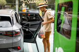 Fototapeta Sypialnia - Man with groceries waiting for electric car to be charged on public charging station near a shopping mall. Man holds mesh bag with fresh vegetables. Sustainable lifestyle concept