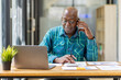 © David - Portrait of Senior African man sitting on laptop at the home office and having a phone conversation with the insurance company.