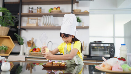 Little toddler asian girl child in apron and chef hat whipped cream decorating preparing homemade cupcakes in home kitchen. A Little girl preparing and decorating homemade cake. Children cooking