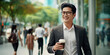 A stylish Asian business man with a cup of coffee walks down the street near the window of a business center.