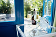 Close up of champagne glasses on transparent table. Bottle of wine and ice cooler. Blue door and garden with green trees on background.