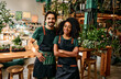 Small flower business. Front view of caucasian man and african american woman in aprons smiling at camera white standing at own shop. Happy couple selling fresh green plants for home decor.