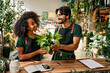 Family business. Caucasian bearded man and african charming woman holding flower plot and smiling to each other. Happy lovely spouse in green apron taking care of plants at own cozy store.