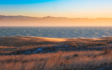  The sea or lake against the backdrop of snowy mountains among the dunes during sunset. Landscape with wind on the Kapchagay reservoir in Kazakhstan Almaty