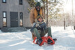 © pressmaster - Happy young African American man in warm winterwear pushing sledge with his son along snow path by their country house