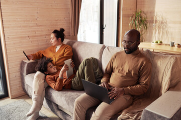 Poster - Cute boy with tablet relaxing on couch between his mother using mobile phone and father typing on laptop keyboard during network