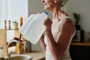 Wall Mural - Cropped shot of senior woman in activewear doing physical exercise with two plastic canisters with water while standing in front of camera