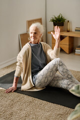 Wall Mural - Senior woman in leggins, t-shirt and cardigan keeping left hand raised and right leg bent in knee while sitting on the floor and practicing yoga