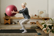© pressmaster - Side view of aged active woman in sportswear holding fitball in front of herself while doing squats on the floor of bedroom in the morning