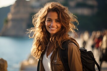 Sticker -  a woman with long hair and a backpack smiles at the camera while standing in front of a body of water with a castle in the backgroufground.