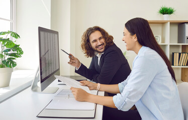 Two happy colleagues having fun at work. Cheerful young man and woman financial accountants sitting at office desk with computer, using business spreadsheets, making funny jokes and laughing