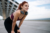 Fototapeta  - Young smiling fit woman in sportswear resting after run wearing headphones. Athletic female listening music taking a breath during a training workout leaning on her knees after exercising.