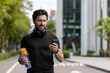 © Tetiana - A young sporty man with headphones is standing on a city street, holding a bottle of water and using a mobile phone