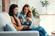 Two happy young women using computer while paying with credit card their vacation sitting on the couch at home.