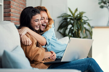 two happy beautiful women doing a video call with laptop while hugging each other sitting on the cou
