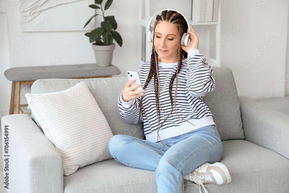 Young woman with dreadlocks in headphones using mobile phone at home