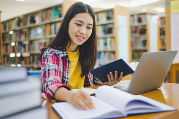 Female college student working on laptop and searching for books to study, make report, find useful information in college room. Concept of reading, learning and intelligence.