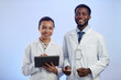 Waist up shot of young African American female laboratory technician holding tablet standing next to Black man scientist while both smiling and looking at camera