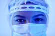 Extreme close up shot of dark-eyed female doctor n blue neon light wearing medical mask and mop cap looking at camera