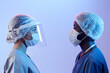 Side view of young female medic in face shield looking at African American male colleague, both wearing masks and sterile hats