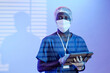 Waist up shot of Black man surgeon in blue light and blinds shadow wearing mask and sterile hat looking at camera and holding tablet