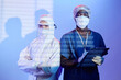 Waist up shot unrecognizable female medic in hazmat suit standing hands crossed by African American male surgeon holding clipboard, both wearing masks and looking at camera