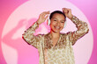 Waist up shot of happy African American girl at bright pink background wearing stylish giraffe print shirt listening to upbeat music with hands in air