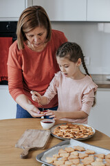 Caucasian woman 60 years old with a girl 8 years old spread jam on cookies in a bright kitchen