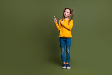 Full length photo of cute schoolgirl with ponytails wear yellow pullover look directing at empty space isolated on khaki color background