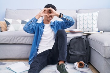 Wall Mural - Young hispanic man sitting on the floor studying for university doing heart shape with hand and fingers smiling looking through sign