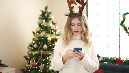 Poster - Young blonde woman using smartphone standing by christmas tree at home