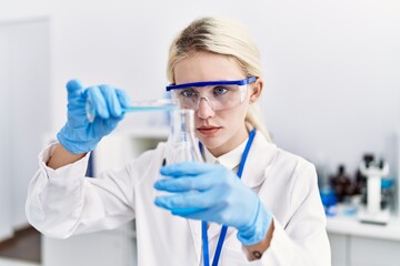 Poster - Young blonde woman scientist pouring liquid on test tube at laboratory