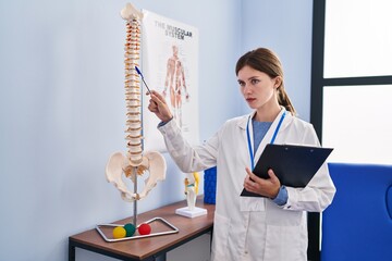 Poster - Young blonde woman pysiotherapist pointing to anatomical model of vertebral column holding clipboard at rehab clinic