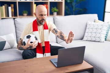 Canvas Print - Hispanic man with tattoos watching football match hooligan holding ball on the laptop with open hand doing stop sign with serious and confident expression, defense gesture