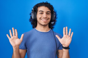 Hispanic man with curly hair listening to music using headphones showing and pointing up with fingers number ten while smiling confident and happy.