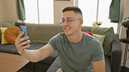 Sticker - Handsome young man taking a selfie indoors with his phone, casually sitting in a modern living room.