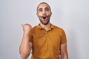 Sticker - Hispanic man with beard standing over white background surprised pointing with hand finger to the side, open mouth amazed expression.