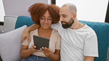 Poster - Beautiful smiling couple sitting together at home, confidently enjoying online bonding over touchpad, radiating joy in their lovely indoor living room relationship